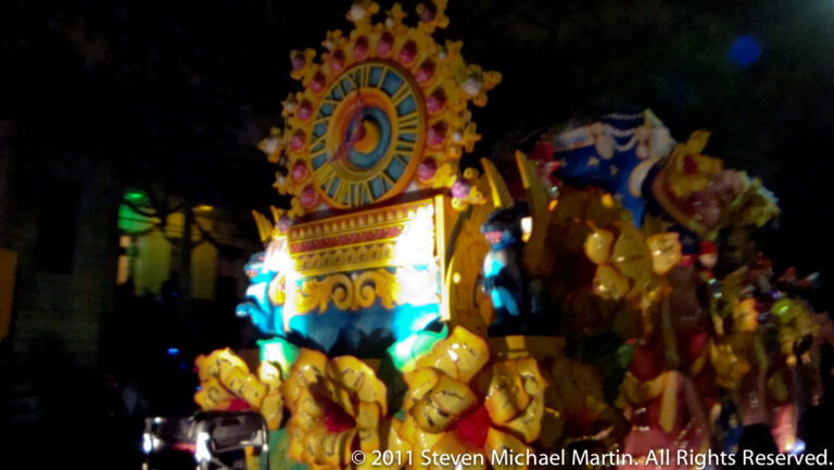 A glowing circular dial crowns a decorated float above rows of small yellow figures and parade participants.