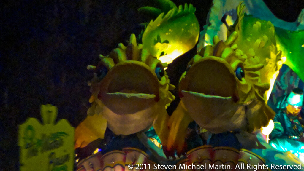 Oversized green frog-like faces rise from a foliage-covered float at night.