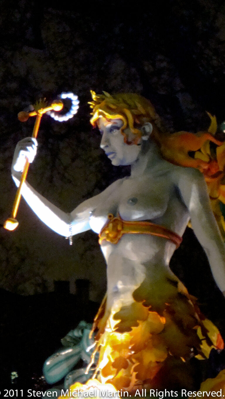 A pale birdlike sculpture spreads its wings across a moving float under green and warm lights.