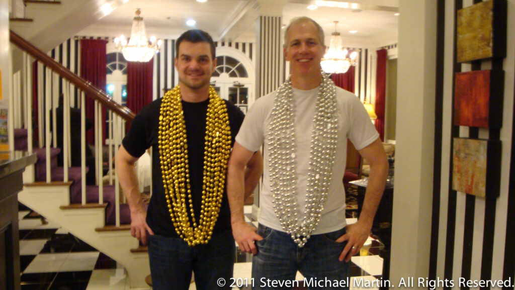 Two men pose in a lobby wearing long strands of Mardi Gras beads.