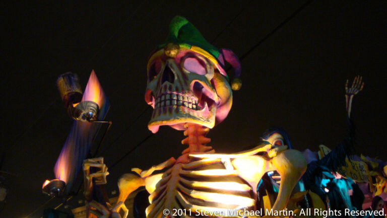 A large skull figure wearing a colorful jester cap glows on a nighttime float.