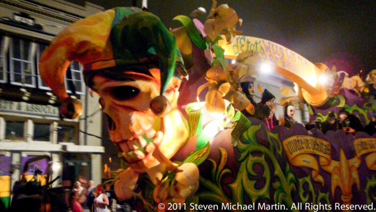 A monumental masked face with a green headdress rises above ornate robes and spectators at night.