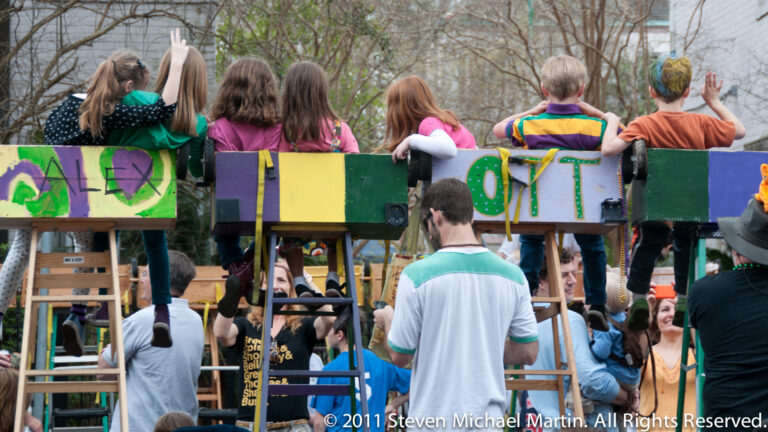 Children ride atop a colorful float while spectators watch from the street below.