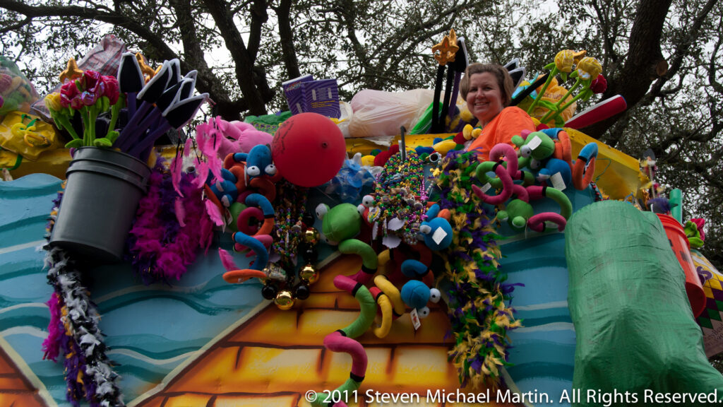 A rider in an orange shirt sits above a crowded aqua float of oversized colorful figures beneath oak trees.
