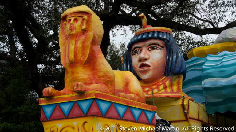 A yellow-orange face and a blue-haired face rise together above a decorated daytime float.