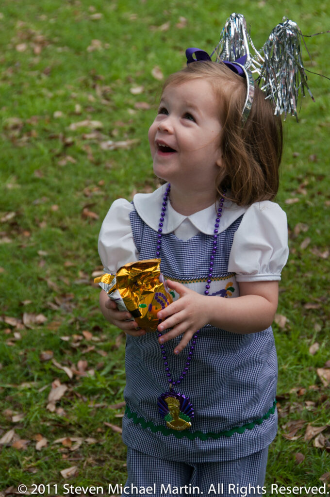 A smiling child wears Mardi Gras beads and metallic headwear while holding a foil-wrapped object.