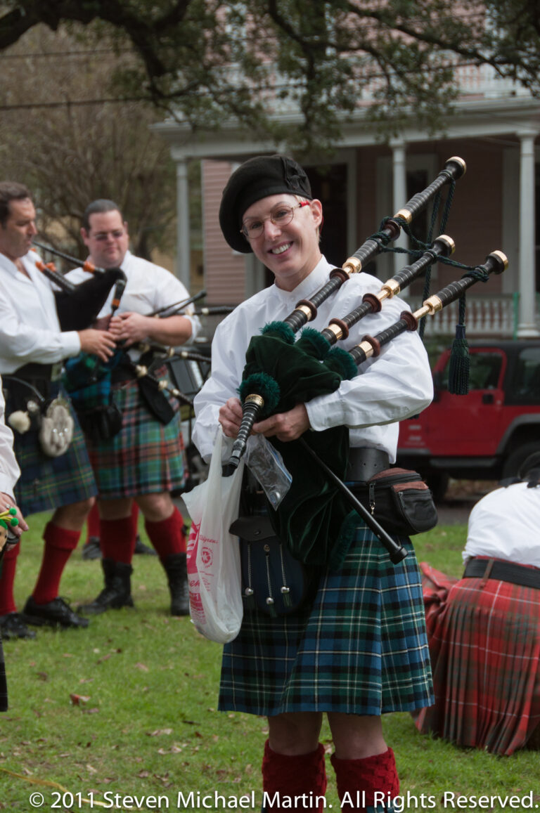 A woman in a tartan kilt plays bagpipes with other pipers following behind.