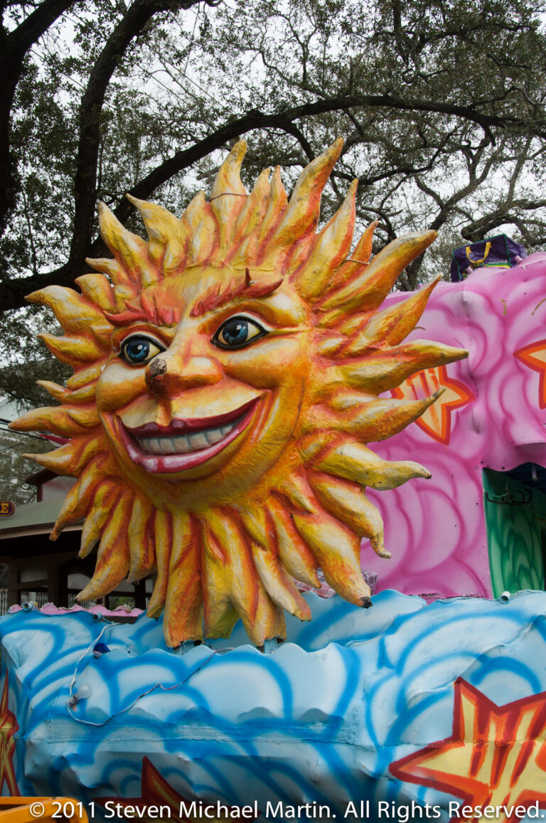 A large smiling sun with radiating rays tops a pink, yellow, and blue parade float.