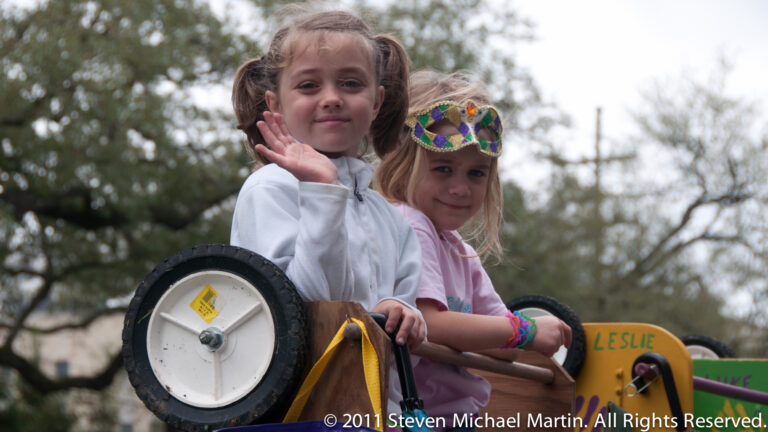 Two girls ride in a small decorated cart; one waves and the other wears a Mardi Gras mask.