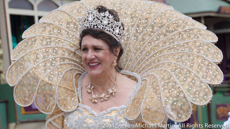 A woman in an ornate crown wears a broad white beaded, feather-like headdress.