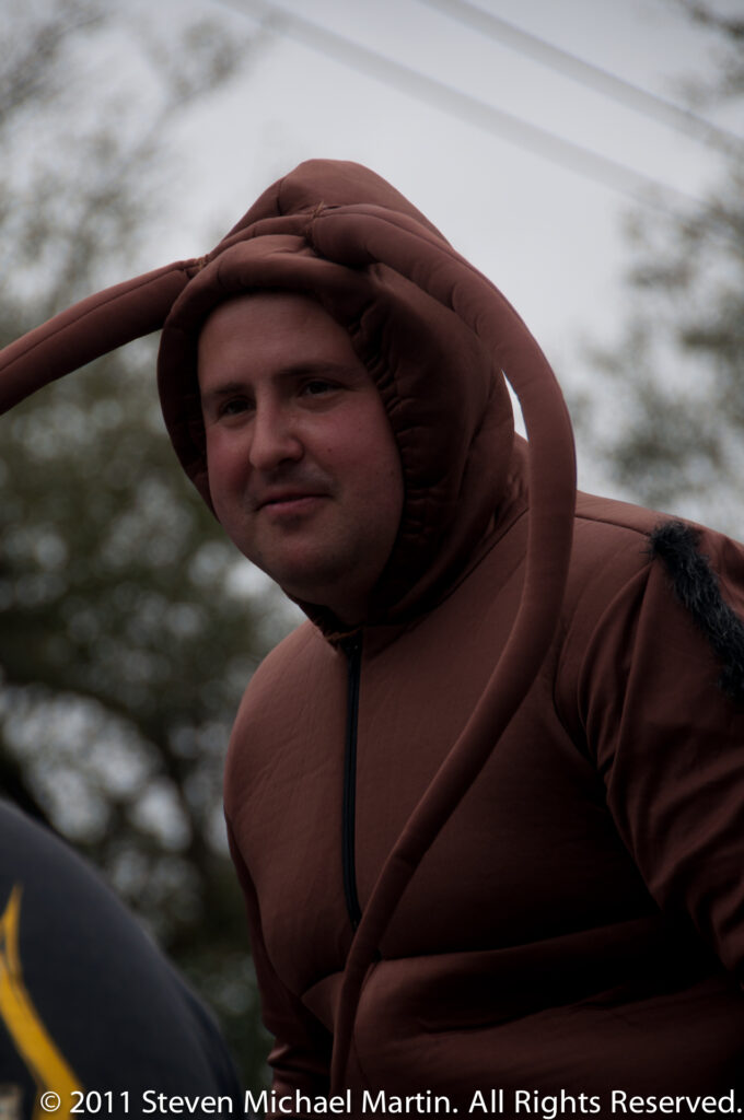 A person wears a brown padded hood with rounded curling forms around the head and shoulders.