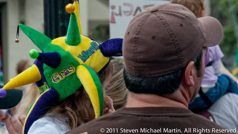 A woman in a green, purple, and gold Mardi Gras jester hat stands among spectators.
