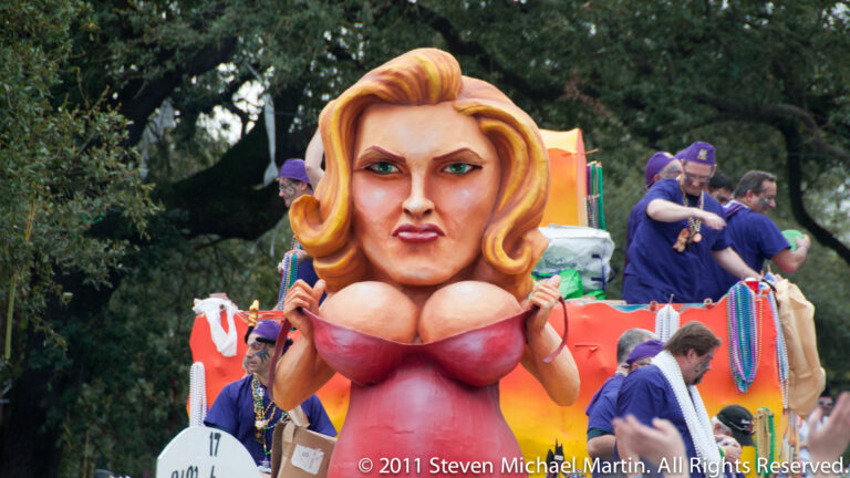 An oversized blonde caricature in a red top rises above costumed workers on a colorful float.