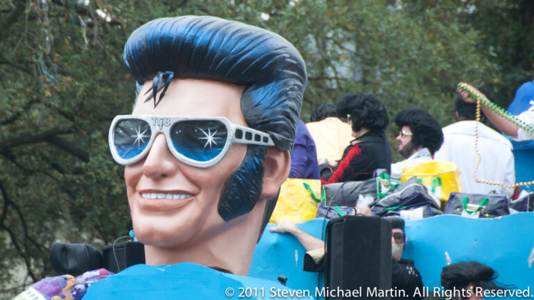 A large pompadour-haired caricature in oversized sunglasses rises above a blue parade float.