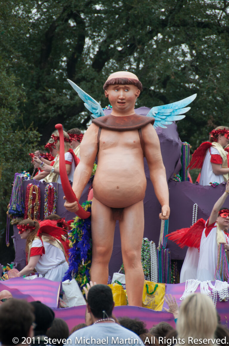 A large winged child figure stands among colorful props and costumed riders on a parade float.