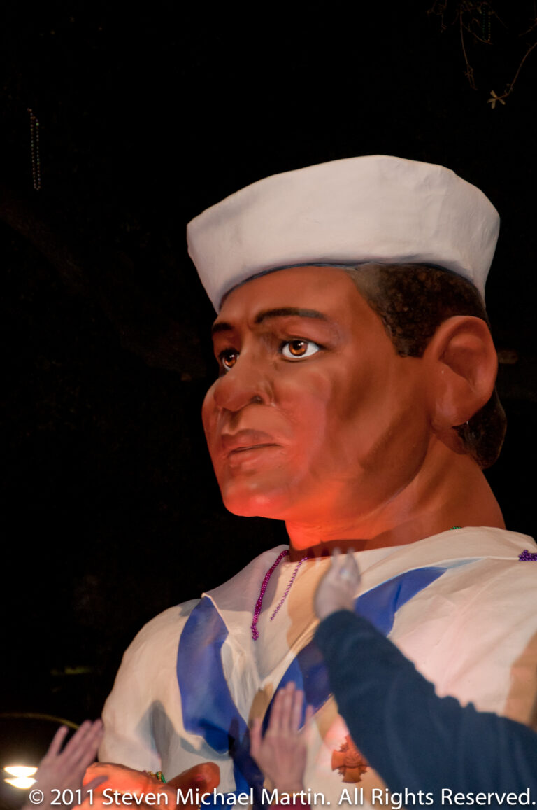 A sculpted male figure in a white sailor cap and blue uniform is lit against a dark float background.