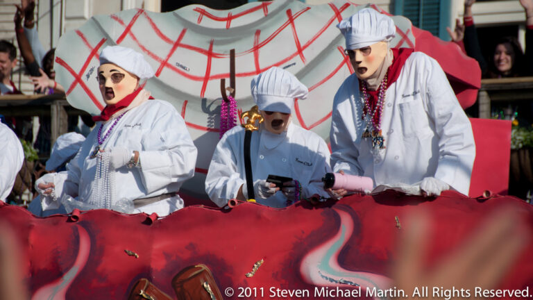 Three performers in white chef uniforms stand behind a red float edge before an oversized clock graphic.