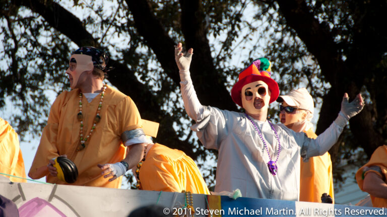 A costumed marcher in a red pointed hat raises both arms beside a participant carrying a small drum.