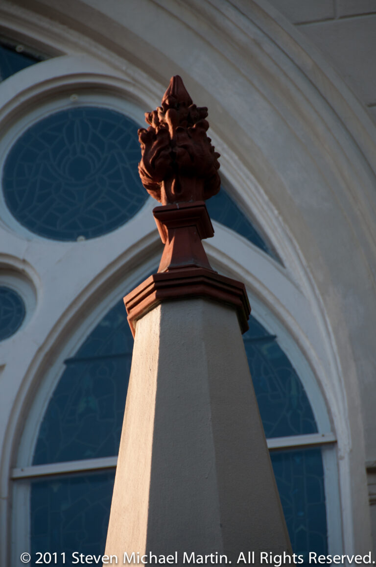 A carved dark finial tops a pale column in front of blue arched windows.