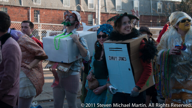 Costumed revelers with wigs and beads carry a cardboard “Davy Jones” sign along a parade route.