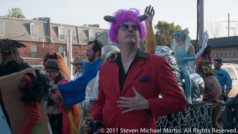 A man in a red suit, purple wig, and tinted glasses stands among costumed revelers.