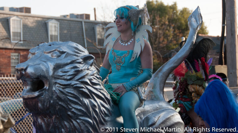 A woman in a turquoise costume rides beside a large silver animal head on a parade float.