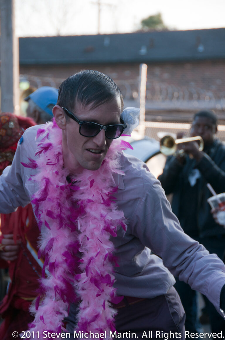 A man in sunglasses and a pink feather boa leans toward the camera beside parade musicians.