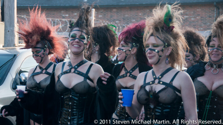 Women in coordinated black costumes and feathered headpieces gather together along a parade street.