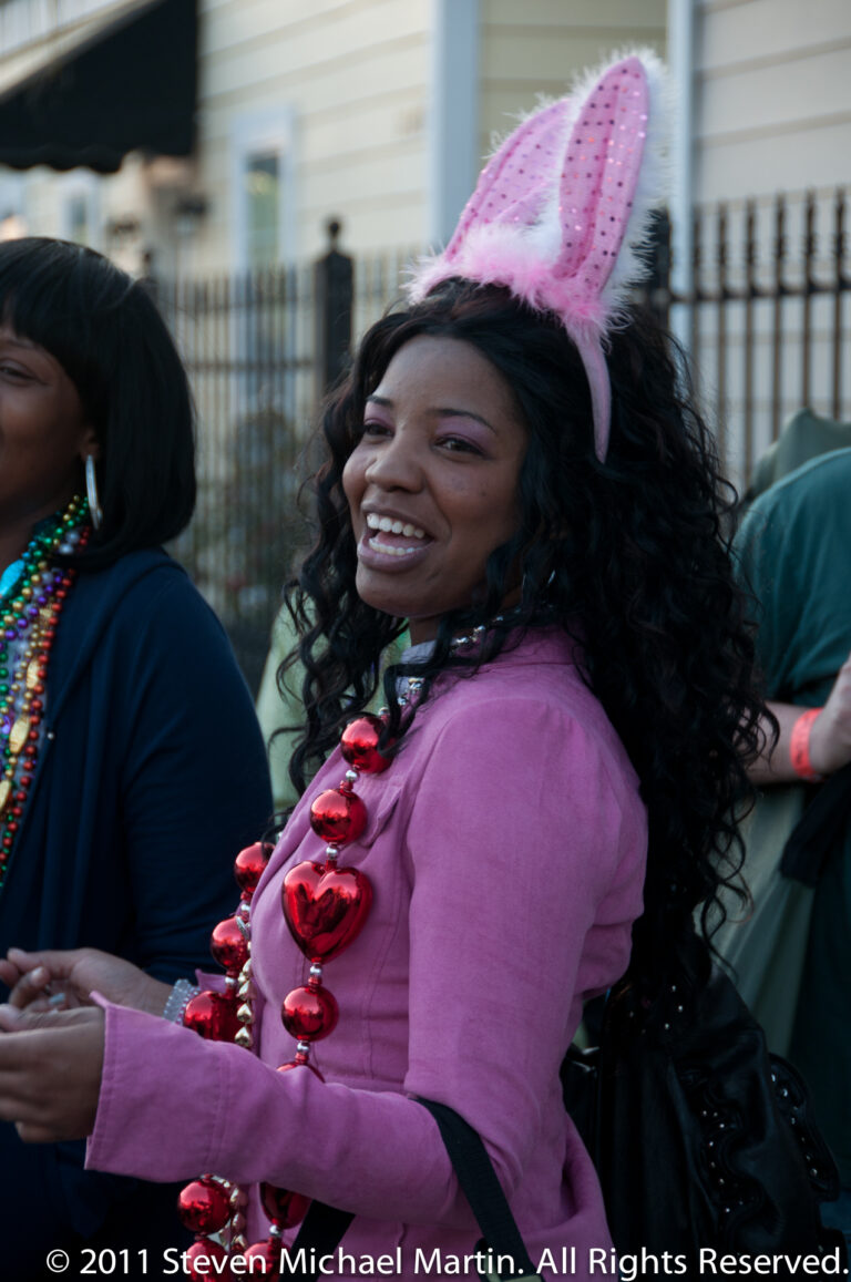 A smiling woman in pink wears a bunny-ear headband and oversized red beads among parade participants.