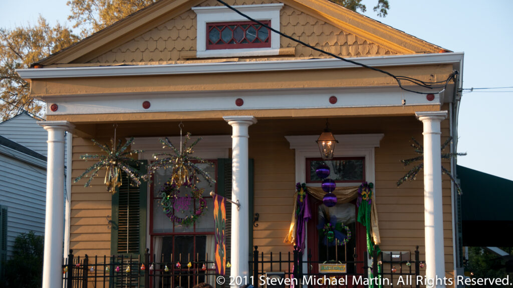 A yellow cottage with a columned gallery displays purple and green Mardi Gras decorations.