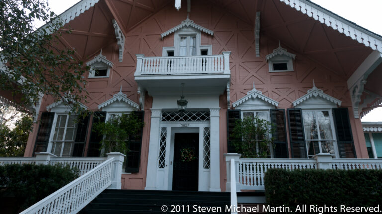 A pink Victorian residence has ornate gables, a small central balcony, and white front steps.