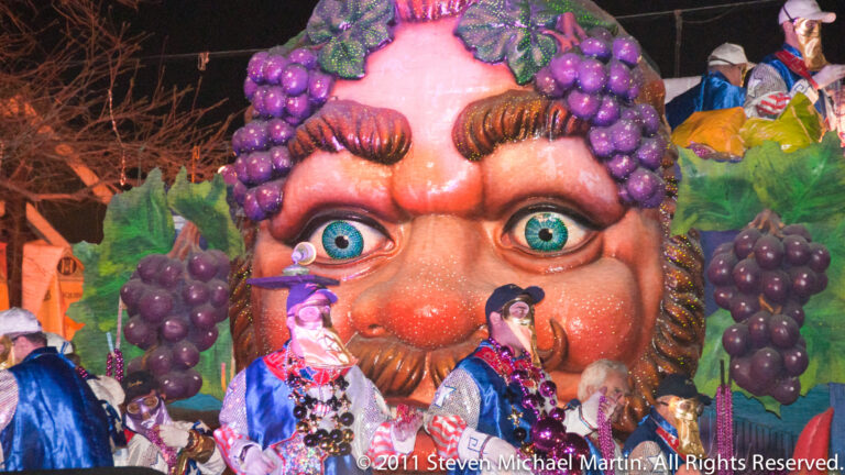 A giant wide-eyed face with a curled moustache is surrounded by purple grapes above smaller float riders.
