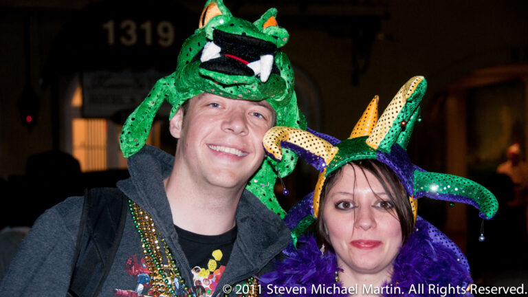 Two smiling revelers wear green alligator hats and Mardi Gras beads in a close portrait.