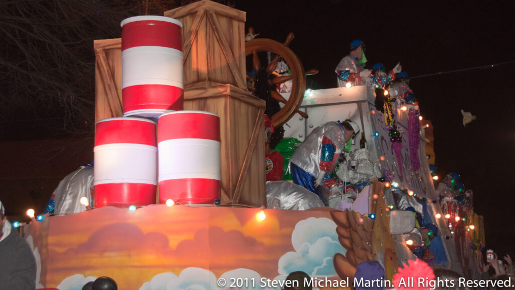A nighttime float carries red-and-white cylinders, a ship’s wheel, colored lights, and costumed riders.