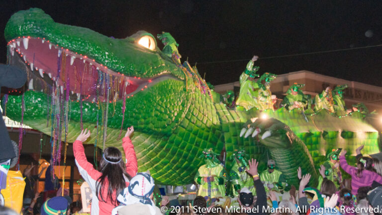 A lit green alligator float carries costumed riders as spectators reach up from the street.