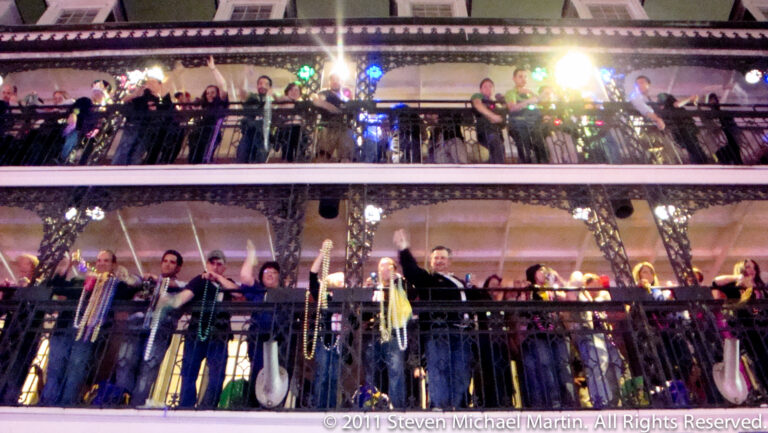 Crowds watch from two stacked wrought-iron balconies at night, with beads hanging from the railings.