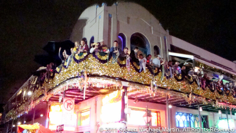 A gold-lit parade float carries costumed riders through a crowded New Orleans street at night.