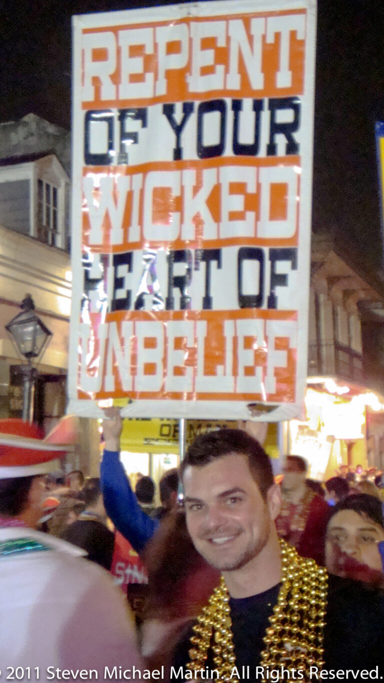 A man wearing gold beads stands beneath a large religious warning sign on a brightly lit street.