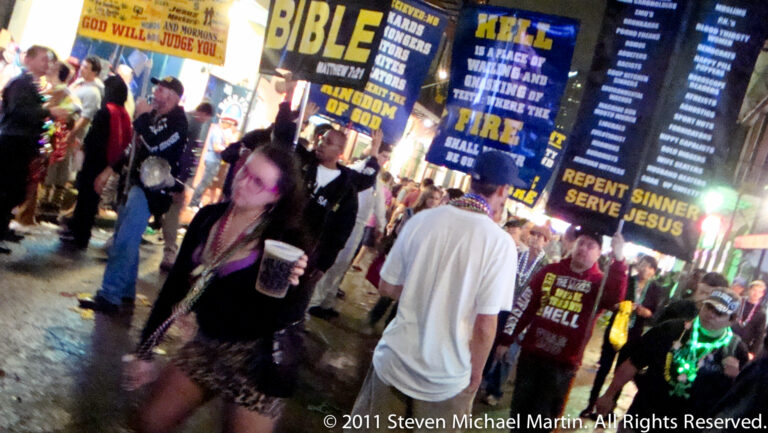 A crowded nighttime street with Mardi Gras beads and tall religious signs about repentance and salvation.