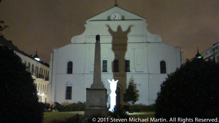A clock-fronted pale building stands at night behind an illuminated statue and a large cast shadow.