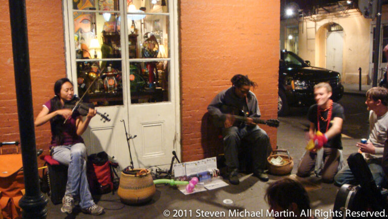 A seated violinist and guitarist perform beside a lit French Quarter storefront with cases and signs on the sidewalk.