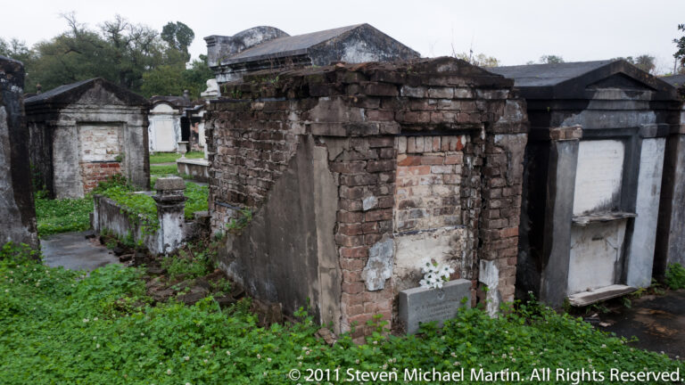 Weathered brick and plaster tombs line a narrow cemetery aisle, with a dark vault in the foreground.