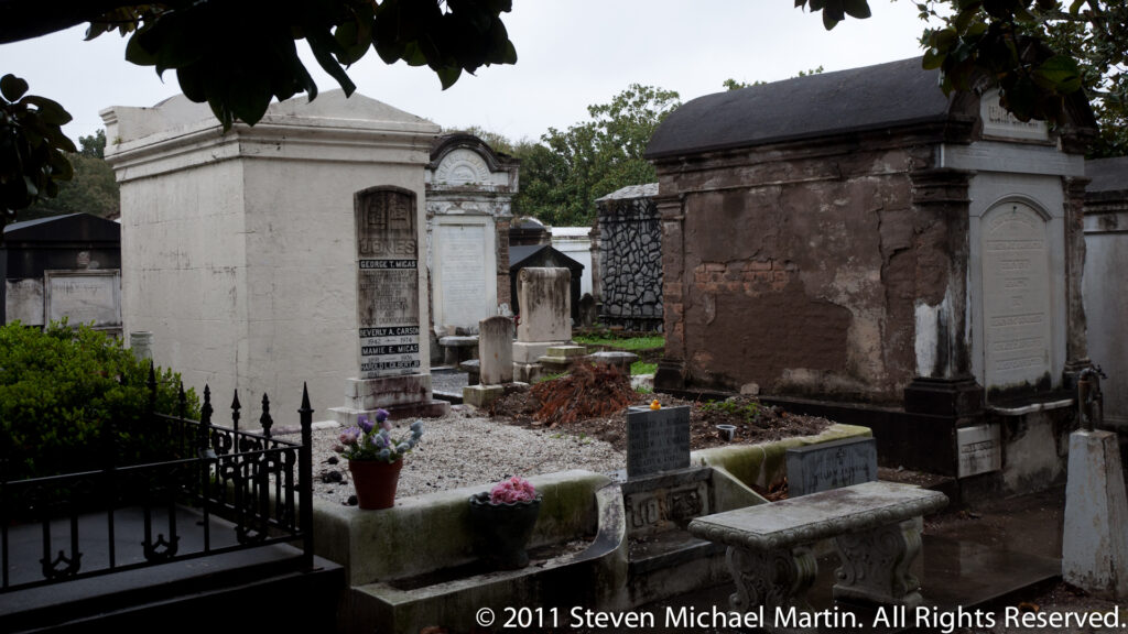 Rows of above-ground tombs and mausoleums surround a narrow cemetery path with flowers and small offerings.
