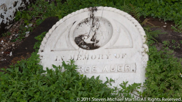 A weathered marble memorial with a carved reclining figure and “In Memory of George Auer” inscription among greenery.