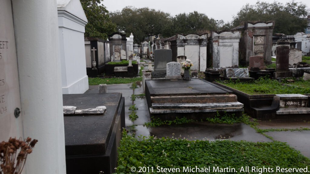 A cemetery aisle recedes between rows of above-ground tombs, with a large white vault close at left.