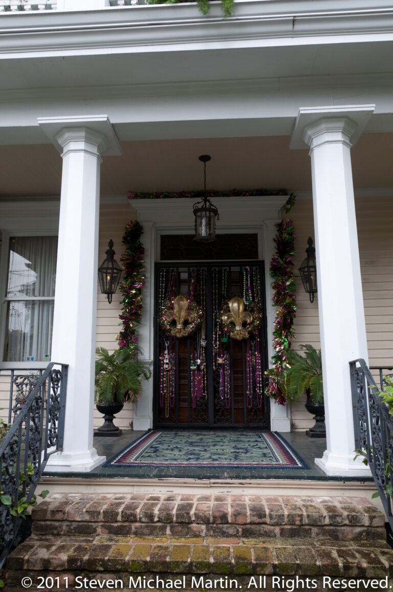 A white columned porch frames dark doors hung with Mardi Gras beads and flanked by potted plants.