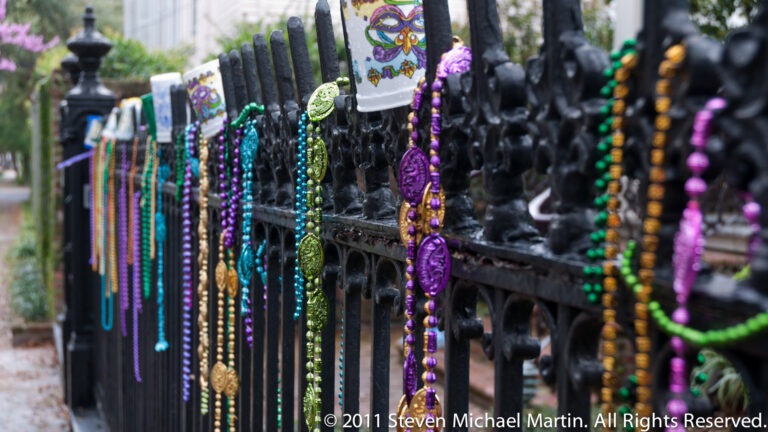 Colorful Mardi Gras bead necklaces and medallions hang from a black wrought-iron fence.