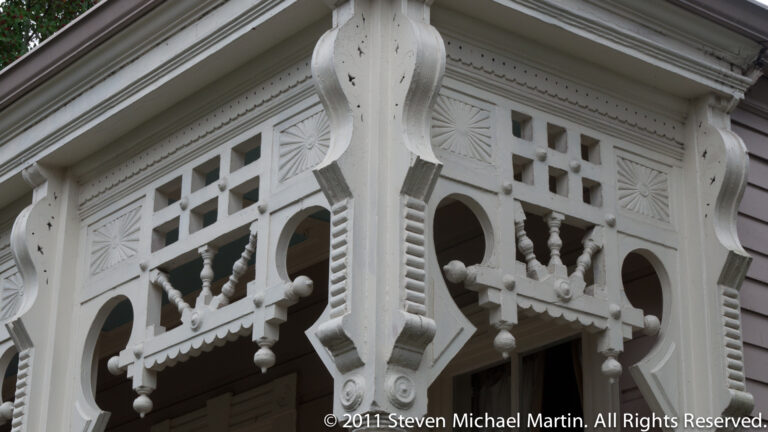 White porch fretwork combines pierced geometric panels, turned balusters, scalloped trim, and a curved bracket.