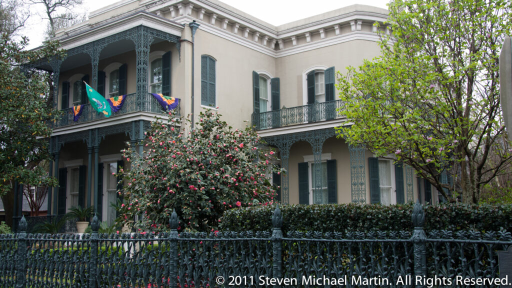 A pale two-story residence with iron galleries and Mardi Gras flags stands behind flowering camellias and a fence.