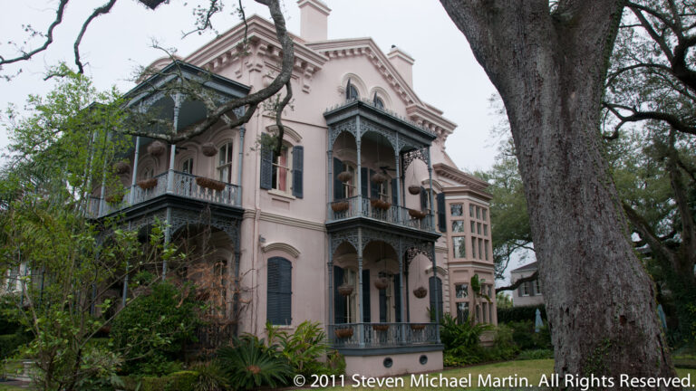 A pale pink residence with dark iron balconies stands beside a massive live oak and dense garden planting.